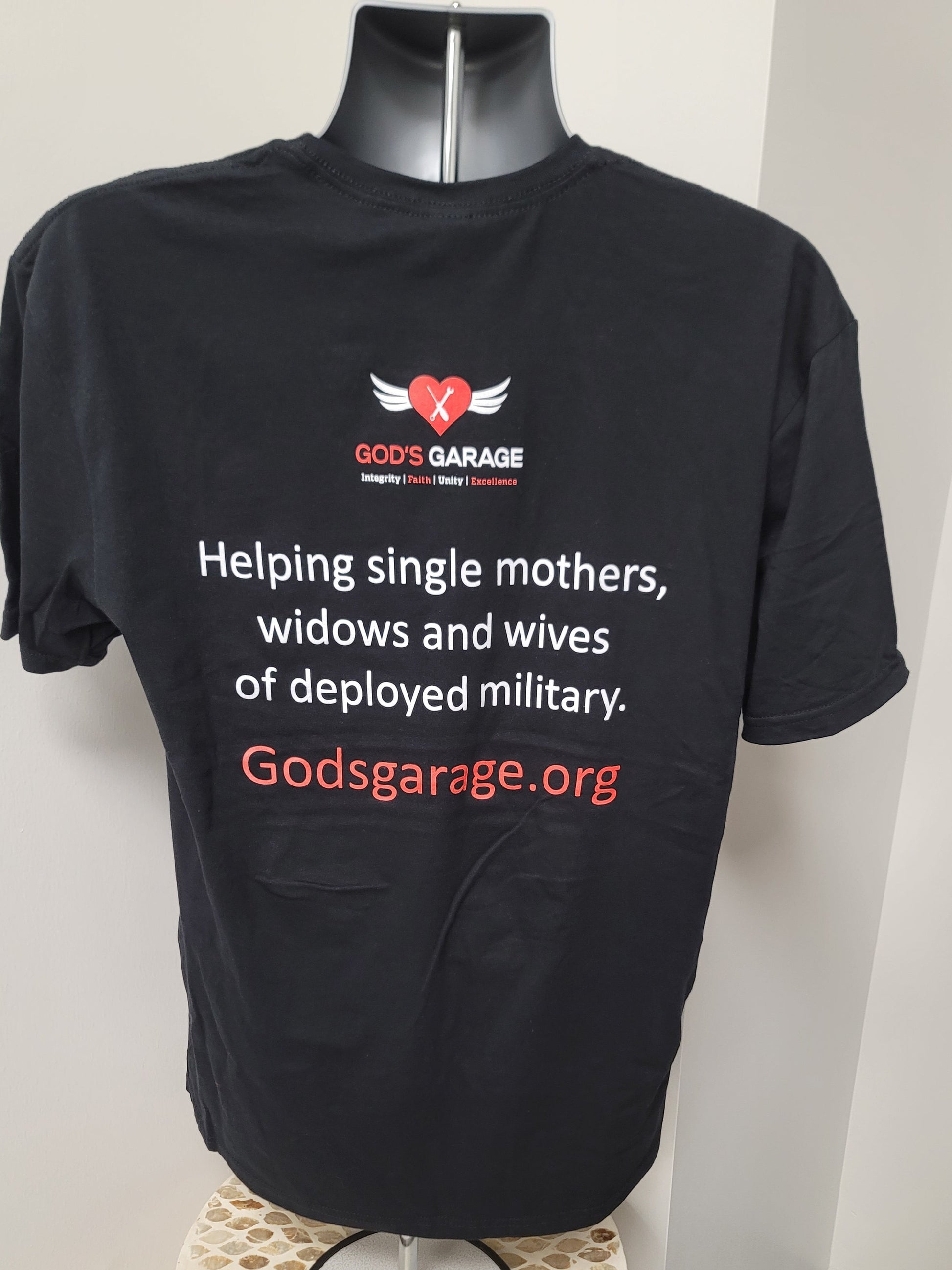 A black short-sleeve t-shirt displayed on a black mannequin torso against a light gray wall. The shirt features a red and white 'God's Garage' winged heart logo on the chest, with white and red text below reading 'Helping single mothers, widows and wives of deployed military.